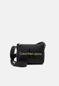 Černá crossbody taška vyrobená z hladké kůže, s embosovaným logem 'CK' a textem 'Calvin Klein Jeans' v žluté barvě. Součástí je nastavitelný popruh.