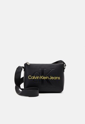 Borsa a tracolla nera in pelle liscia, con logo 'CK' impresso e testo 'Calvin Klein Jeans' in giallo. Inclusa tracolla regolabile.