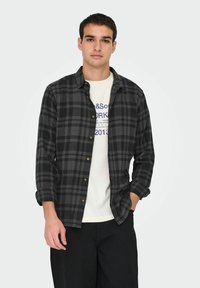 Zwart en grijs geruit overhemd met lange mouwen, knoopsluiting aan de voorkant, twee borstzakken, gedragen over een wit graphic T-shirt.