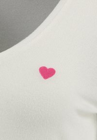 Top in tessuto bianco con un'emblema a cuore ricamato rosa vicino al collo. Texture morbida con un design a scollo rotondo.
