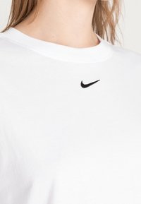 Bílý bavlněný tričko s kulatým výstřihem, s malým černým logem Nike vyšitým na hrudi. Hladký, lehký materiál.