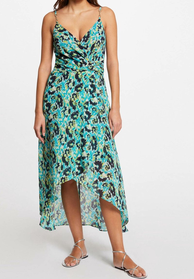 Femme portant une robe portefeuille sans manches à fleurs bleu, vert et noir avec un ourlet asymétrique et des sandales argentées à brides.