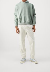 Sudadera de algodón verde claro con bolsillo canguro, combinada con pantalones de algodón blancos y zapatillas deportivas azules con detalles blancos.