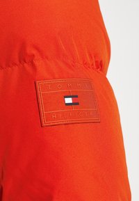 Röd tyg med en rektangulär Tommy Hilfiger-logotyp i patchform, som har vita, röda och blå ränder samt präglat varumärkesnamn.