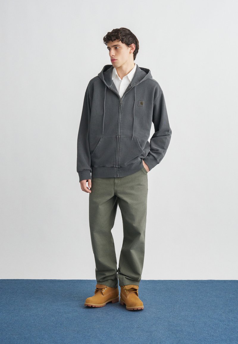 Young man wearing a dark gray zip-up hoodie, white shirt, olive green pants, and tan work boots standing on blue carpet.