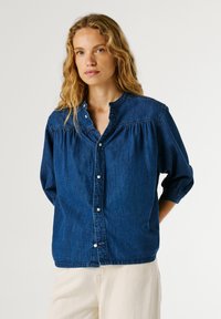 Femme aux cheveux ondulés portant une chemise en jean bleue boutonnée avec des manches trois-quarts et un pantalon de couleur claire sur un fond uni.