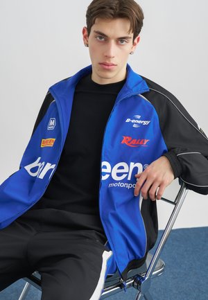 Νέος άνδρας με ανοιχτόχρωμα μάτια, φορώντας ένα μπλε και μαύρο μπουφάν μηχανοκίνητου αθλητισμού και μαύρο παντελόνι, καθισμένος σε μεταλλική καρέκλα σε απλό φόντο.