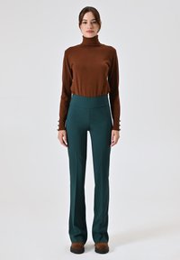 Maglione a collo alto marrone e pantaloni teal a vita alta. I pantaloni hanno gamba larga. Le scarpe marroni completano l'outfit. Sfondo piatto.