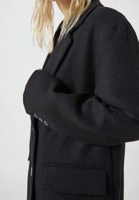 Černý blazer z vlněné směsi s klopovým límcem, jednou náprsní kapsou a knoflíkovým zapínáním na třech knoflících na manžetách. Viditelná texturovaná tkanina.