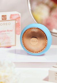Foreo-enhet med ett turkos silikonutsida och en texturerad roséguldfinish, uppställd tillsammans med en produktlåda på en vit yta.