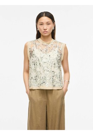 Kvinde med glat mørkt hår iført ærmeløs beige top med sølv palietter og løse lysebrune bukser, stående med hænderne i lommerne.