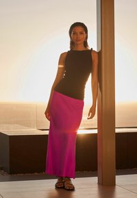 Top nero senza maniche e gonna lunga fucsia con una texture liscia. La modella si trova accanto a una colonna, con la luce solare che crea un alone luminoso dietro di lei.