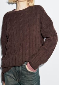 Maglione marrone a maglia con collo rotondo e polsini a costine, indossato su una camicia a maniche lunghe rosa, abbinato a jeans di denim scuri.