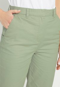 Lichtgroene broek met een gladde textuur, voorzien van zijvakken en een knoopsluiting in de taille. De stof lijkt duurzaam en ademend.