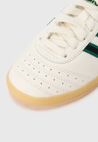 Biały sneaker z gładką, skórzaną cholewką. Posiada zielone i czarne paski, perforacje dla wentylacji oraz gumową podeszwę.