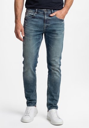 Man die slim-fit vervaagde blauwe spijkerbroek en witte sneakers draagt, staand met één hand in zijn zak en de andere ontspannen langs zijn zijde.