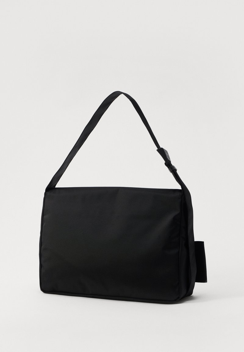 Sac messager noir en nylon lisse, présentant une forme rectangulaire plate, une bandoulière unique et un design minimaliste sans accents métalliques.