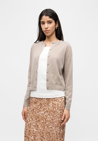 Μπεζ εφαρμοστό ζακέτα, απαλή υφή, με κουμπιά μπροστά και στρογγυλή λαιμόκοψη. Φορεμένο πάνω από λευκό τοπ και φούστα σε ροζ χρυσό με παγιέτες.