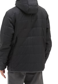 Mężczyzna w czarnej, pikowanej kurtce z kapturem i szarych spodniach, pokazany od tyłu na białym tle.