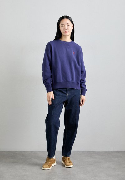 Sudadera morada con cuello redondo y puños acanalados, acompañada de jeans oscuros holgados y zapatillas de ante marrón. Sin patrones visibles.