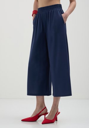 Personne portant un pantalon large cropped bleu marine, des chaussures à talons hauts rouges avec bride arrière et un bracelet rouge, debout les mains dans les poches sur un sol blanc.