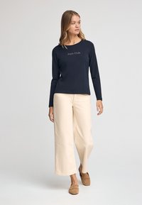 Marineblaue Langarm-T-Shirt mit "Polo Club"-Schriftzug, kombiniert mit cremefarbenen weit geschnittenen Hosen und braunen Schlupfschuhen auf einem grauen Hintergrund.