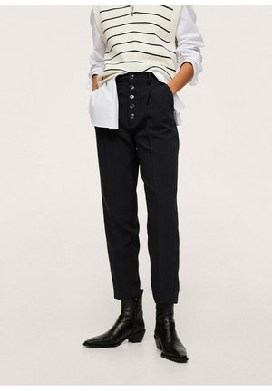 Osoba nosząca czarne, wysokie spodnie zapinane na guziki, czarne botki, białą koszulę z podwiniętymi rękawami oraz pasiasty bezrękawnik.