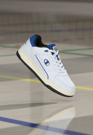Weißer und blauer Sportschuh mit Schnürsenkeln, schwarzer Sohle und Logo an der Seite und Zunge, in der Luft über einem Hallenboden mit Spielfeldlinien gezeigt.