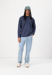 Navy long-sleeve shirt with logo, light blue jeans, and green cap. White shoes with red accents. Casual outfit with relaxed fit.