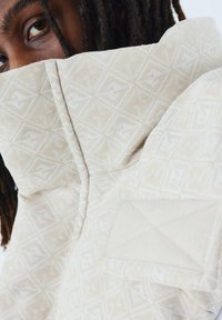 Beige gestreepte jas met een hoge kraag, textuurstof en een driehoekige patch. Subtiel geometrisch ontwerp in een licht kleurenschema.
