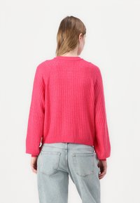 Stickad tröja i knallrosa, med ribbat mönster, rund halsringning och avslappnad passform med långa ärmar; tillsammans med ljusblå jeans.