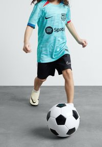 Turkosfärgad FC Barcelona-tröja med röda och blå accenter, svarta Nike-shorts och gula fotbollsskor som sparkar en svartvit fotboll.