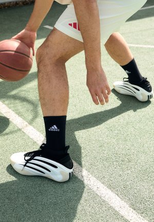 Personne portant des chaussures de basketball blanches Adidas et des chaussettes noires dribblant un ballon de basket sur un terrain extérieur avec une surface verte.