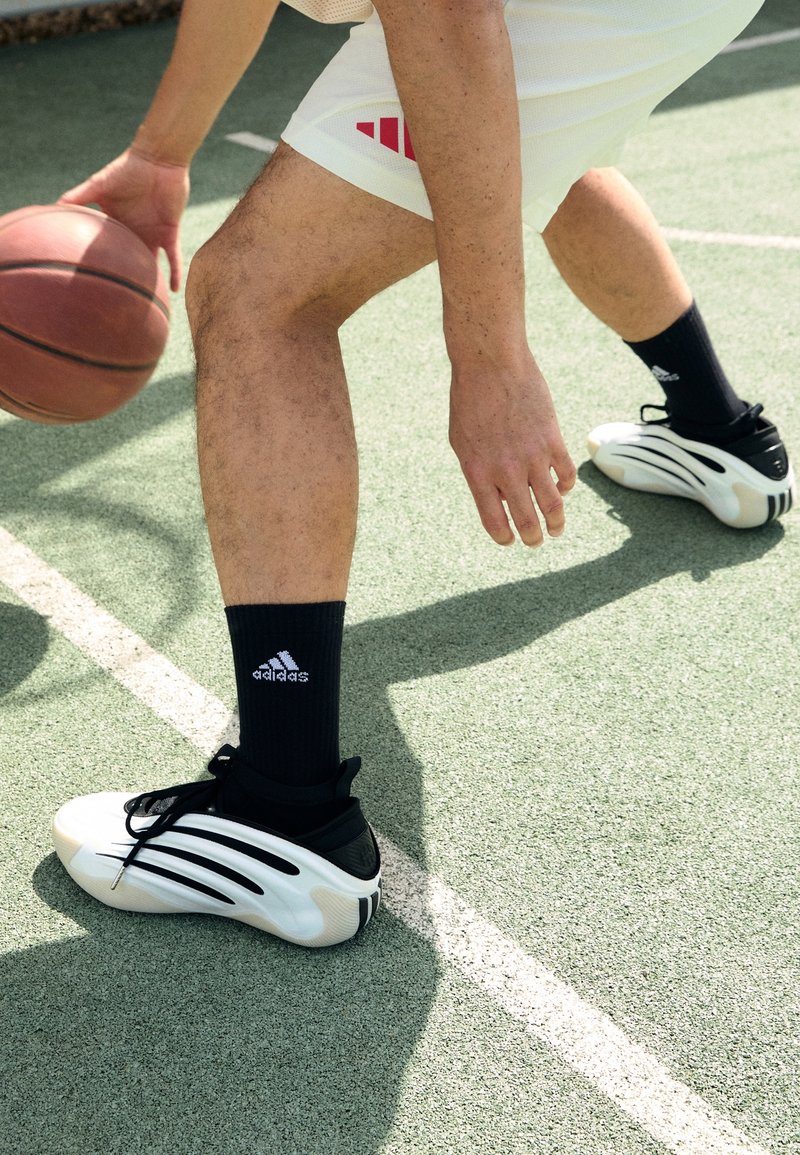 Personne portant des chaussures de basketball blanches Adidas et des chaussettes noires dribblant un ballon de basket sur un terrain extérieur avec une surface verte.