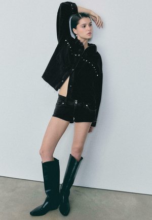 Młoda kobieta w czarnej kurtce z ćwiekami, czarnych szortach i czarnych kozakach do kolan pozuje z uniesioną jedną ręką na tle jednolitej ściany.