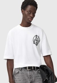 Valkoinen puuvillapaita rentoa mitoitusta, pyöreä pääntie ja lyhyet hihat. Vasemmassa rinnassa on musta graafinen logo.