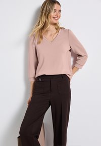 Frau trägt eine hellrosa V-Ausschnitt-Bluse und eine dunkelbraune weitgeschnittene Hose, steht gegen eine weiße Wand und schaut mit einem Lächeln zur Seite.