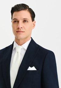 Granatowa marynarka z klapami w szpic, o fakturowanym materiale, biała koszula i kremowy krawat. Złożona biała poszetka znajduje się w piersiowej kieszeni.