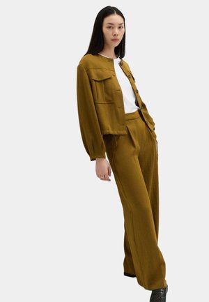 Kvinde iført sennepsfarvet knappet jakke og matchende bukser med vidde, kombineret med en hvid top og sorte sko, stående foran en hvid baggrund.