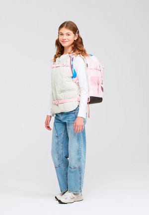 Girl standing wearing blue jeans, white long sleeve shirt, padded vest, and pink backpack with chest and waist straps fastened.