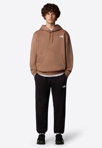 Bruine hoodie met een voorzak en trekkoordkap; zwarte sweatpants met logo; gecombineerd met zwarte schoenen. Eenvoudige en casual sportoutfit.