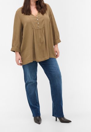 Kvinde iført en olivengrøn bluse med lange ærmer og knapper, blå jeans og sorte spidsede støvler med hæl, stående mod en hvid baggrund.