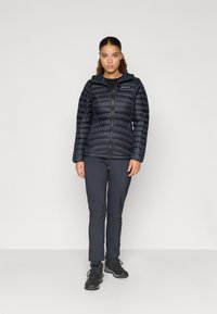 Czarna kurtka puchowa z poziomym pikowaniem, zapinana na zamek, z kapturem; zestawiona z ciemnymi spodniami i czarnymi butami trekkingowymi na neutralnym tle.