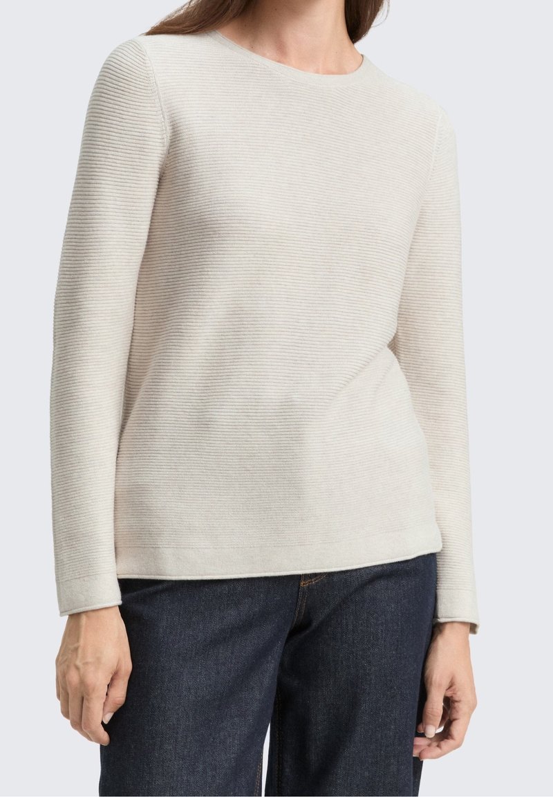 Pull côtelé beige clair à manches longues porté avec un jean bleu foncé, montré sur une personne de la nuque aux hanches sur un fond uni.