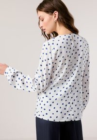 White blouse with navy blue polka dots, long sleeves, rounded neckline, and a button detail at the back. Smooth fabric with a relaxed fit.