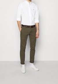 Bílá košile s dlouhým rukávem a knoflíky, olivově zelené slim-fit kalhoty, hnědý opasek a bílé tenisky, stojící na světle šedém pozadí.