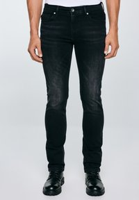 Férfi fekete, slim fit farmernadrágban és fekete fűzős bakancsban, sima világosszürke háttér előtt áll.