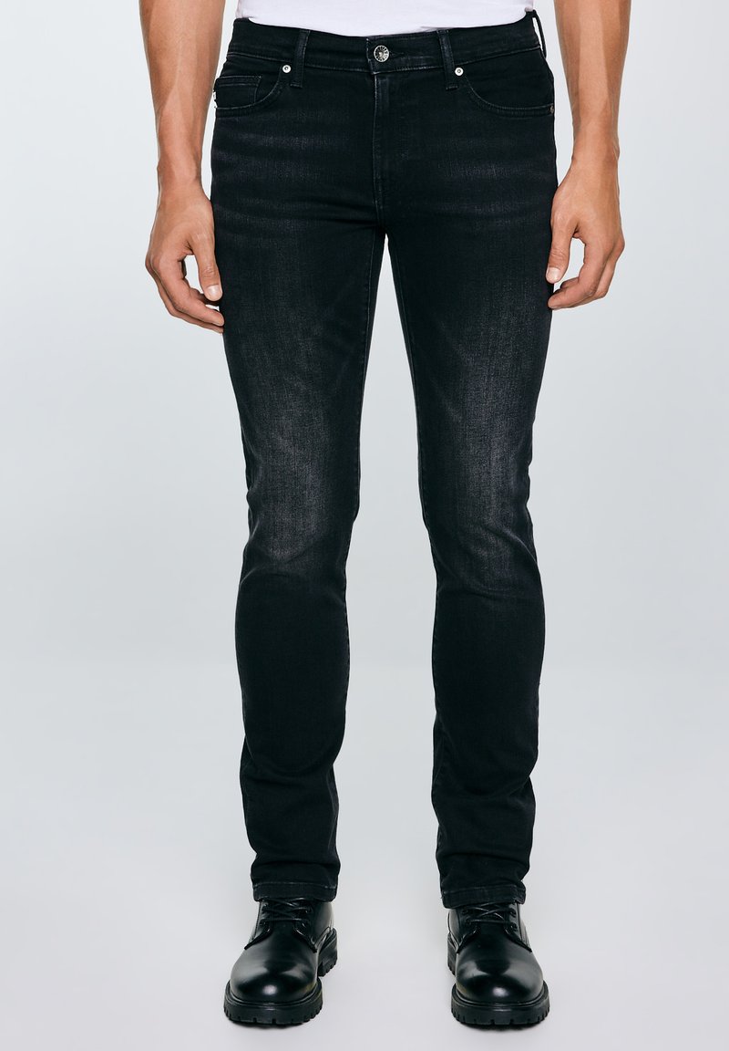 Férfi fekete, slim fit farmernadrágban és fekete fűzős bakancsban, sima világosszürke háttér előtt áll.