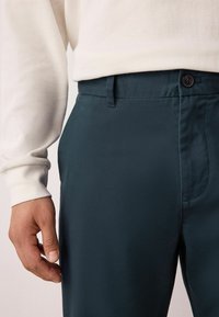 Pantalon bleu foncé en tissu lisse, avec une taille plate, des poches latérales et une fermeture à bouton. Associé à un haut blanc à manches longues.