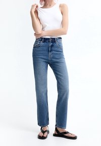 Jeans de mezclilla azul de talle alto con pierna recta, que cuentan con cinco bolsillos y un sutil efecto desgastado, combinados con un top blanco sin mangas y sandalias negras.